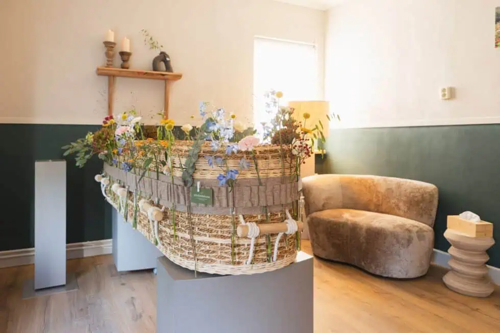 Rieten uitvaartkist met veldbloemen naast een comfortabele stoel
